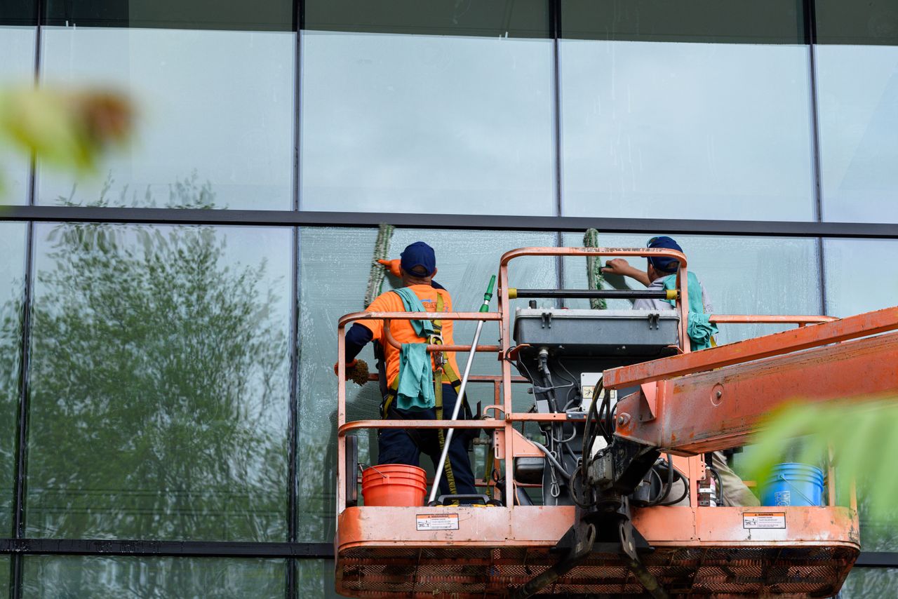 Deux travailleurs nettoient les fenêtres d'une façade en verre à l'aide d'une nacelle élévatrice, ce qui améliore l'esthétique du bâtiment.